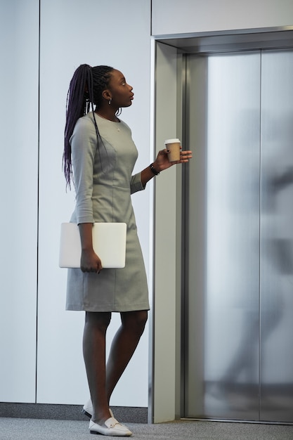 Pionowy portret pełnej długości afroamerykańskiej bizneswoman czekającej na windę w budynku biurowym i trzymającej filiżankę kawy