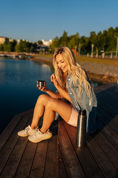 Pionowy portret roześmianej blondynki młodej kobiety trzymającej kubek z gorącą kawą z termosu siedzącego na plaży przy molo w letni słoneczny poranek