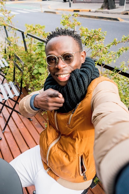 Pionowy portret selfie młody afrykański mężczyzna siedzący na zewnątrz uśmiechający się i wskazujący na kamerę