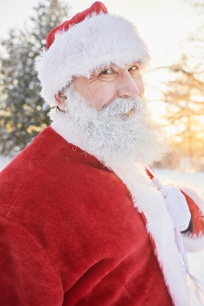 Pionowy portret uśmiechniętego Świętego Mikołaja patrzącego na kamerę na zewnątrz w zimowym lesie i niosącego sa