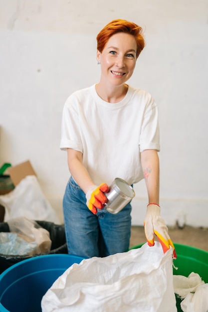 Zdjęcie pionowy portret wesołej pracownicy w lateksowych rękawiczkach patrzącej na kamerę i sortującej wyrzucone metalowe puszki w nowoczesnym prywatnym zakładzie recyklingu odpadów