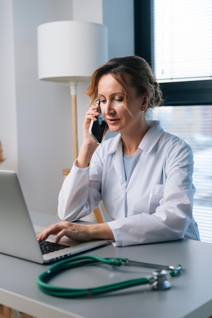 Pionowy portret zajęty lekarka w białym fartuchu, pisanie na laptopie i rozmowa na smartfonie, siedząc przy biurku w klinice lekarskiej na tle okna