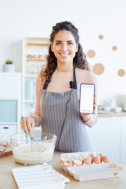 Pionowy średni portret pięknej kobiety rasy kaukaskiej stojącej w nowoczesnej kuchni trzymając smartfon demonstrując nową aplikację