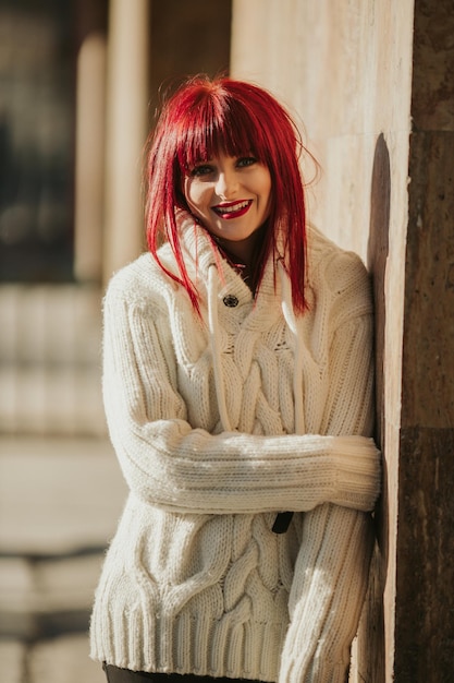 Pionowy zdjęcie młodej uśmiechniętej kobiety z czerwonymi włosami pozującej w ciepłym białym swetrze