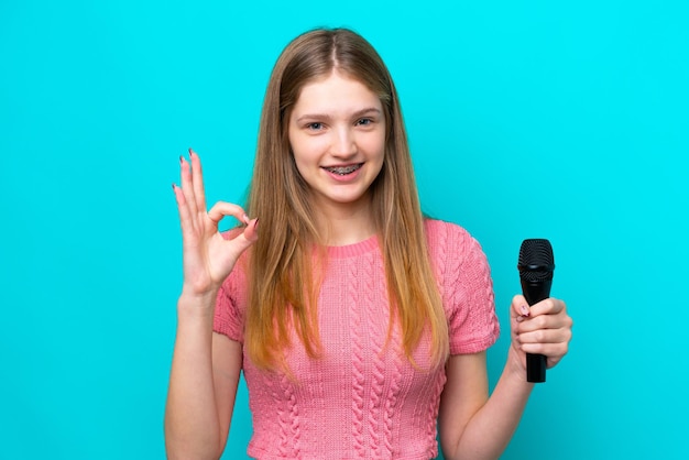 Piosenkarka Rosyjska dziewczyna podnosząca mikrofon na białym tle na niebieskim tle pokazująca znak ok palcami