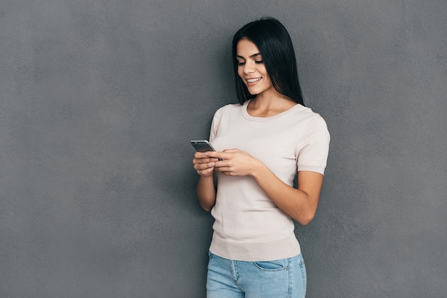 Pisanie wiadomości do przyjaciela. Atrakcyjna młoda kobieta trzymająca inteligentny telefon i patrząca na niego z uśmiechem, stojąc na szarym tle