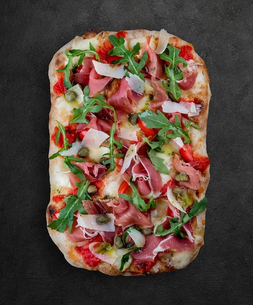 Pizza Sorrentina z prosciutto rukolą kaparami pelati sos pesto rzymska pizza prostokątna na ciemnym tle