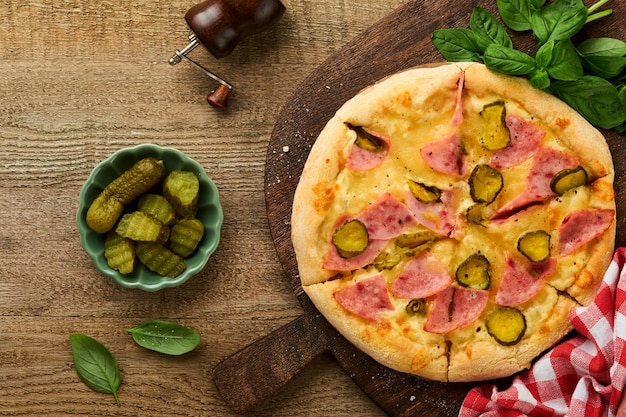 Pizza Tradycyjna pizza na bekonie z szynką, grzybami, marynowanym ogórkiem i serem oraz składnikami do gotowania, pomidorami, bazylią na drewnianym stole, włoska, tradycyjna kuchnia, widok z góry, makieta