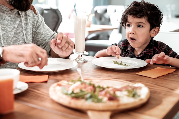 Pizza z ojcem. Ciemnooki, rozpromieniony, kręcony chłopiec czuje się wyjątkowo pozytywnie i szczęśliwie jedząc pizzę z ojcem