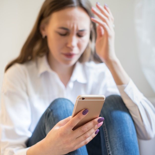 Płacząca kobieta w depresji patrząc na telefon dostaje złe wieści, siedzi, skupia się na smartfonie.