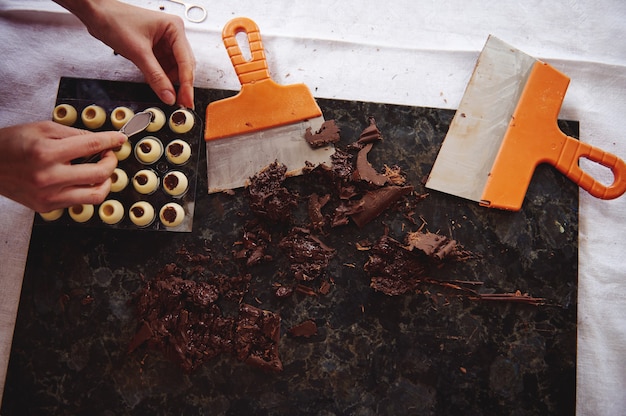 Płaska kompozycja z rękami cukiernika wkładającego nadzienie czekoladowe do kulistych foremek z białej czekolady. Hartowana masa czekoladowa i szpachlówki leżące na marmurowej powierzchni