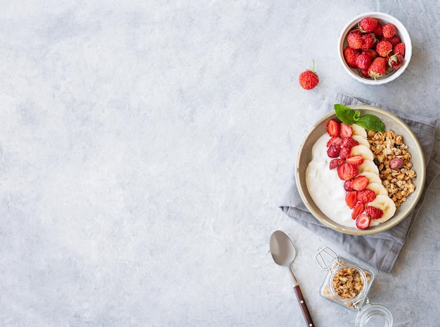 Płaska warstwa naturalnego jogurtu z truskawką granola i bananem w misce na jasnym tle z świeżymi jagodami i łyżką