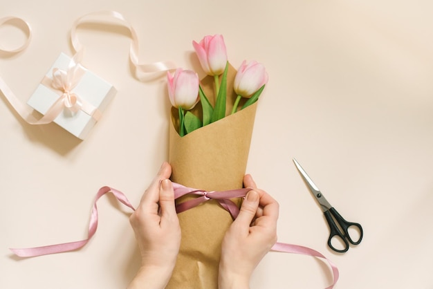 Płaskie kobiece dłonie zawiązują satynową wstążką łuk na prosty bukiet kwiatów świeżych różowych tulipanów w papierze rzemieślniczym na beżowym stole.