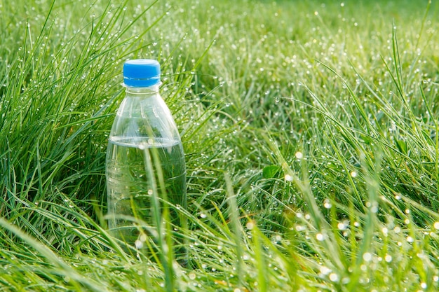 Plastikowa przezroczysta butelka wody mineralnej w zielonej trawie z kroplami rosy wczesnym rankiem. Selektywne skupienie się na butelce.