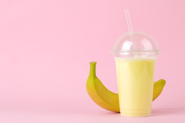 Plastikowa szklanka jednorazowa z bananowym koktajlem mlecznym i świeżym bananem na jasnym, modnym różowym stole. Robienie koktajlu mlecznego.