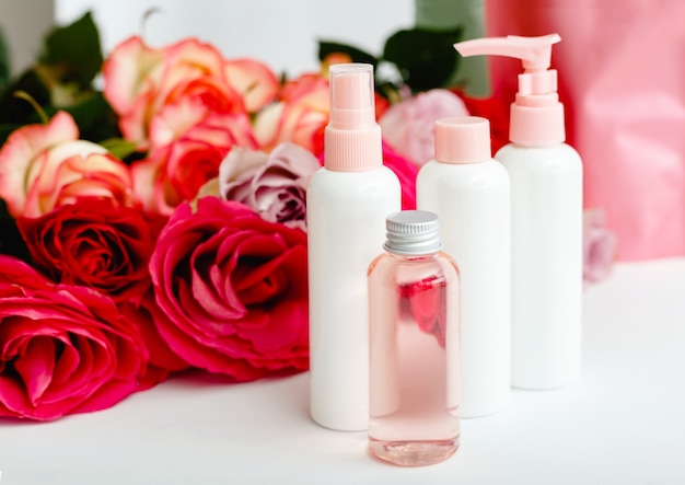 Plastikowe butelki kosmetyczne, serum, mydło, olej na białym stole kwiatowym. Kwiat czerwony różowy róż naturalny organiczny produkt kosmetyczny. Spa, pielęgnacja skóry, kąpiele na ciało. Zestaw kosmetyków w kolorze różowym z różą.