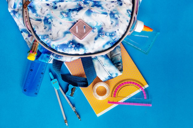 Plecak z kolorowymi ołówkami, notatnikami, linijką, flamastrami na niebieskim tle