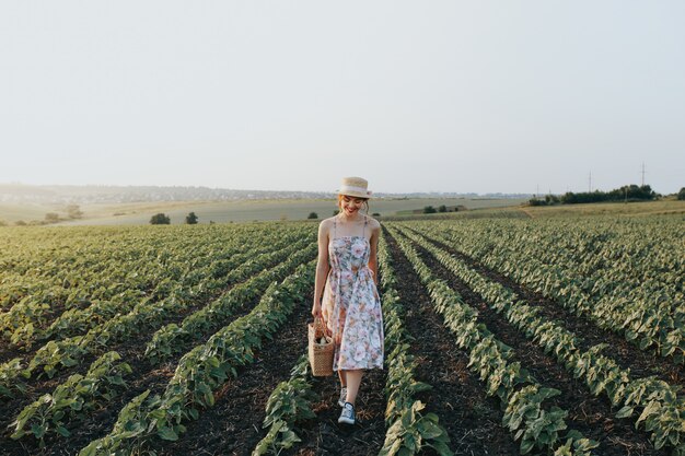Plenerowy lato portret nastoletnia dziewczyna z koszykowymi truskawkami, słomiany kapelusz. Dziewczyna na wiejskiej drodze, widok z tyłu. Natury tło, wiejski krajobraz, zielona łąka, kraju styl