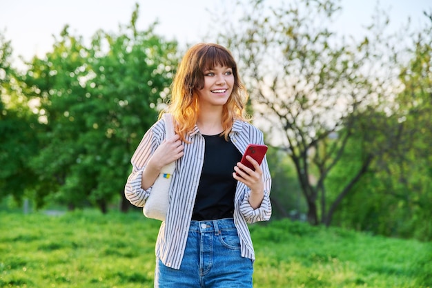 Plenerowy portret modnej młodej rudowłosej dziewczyny ze smartfonem w ręku zielone drzewa na tle nieba Uśmiechnięta atrakcyjna nastoletnia studentka Koncepcja wypoczynku młodzieży uroda moda styl życia