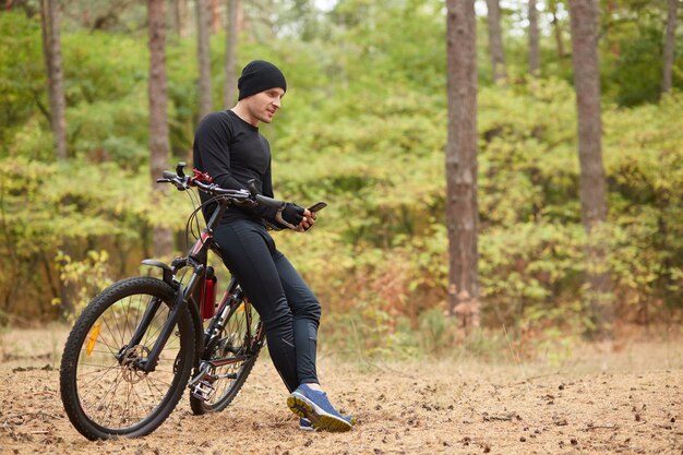 Plenerowy strzał uśmiechnięta silna sportowiec pozycja w jesień lesie, opierający się na jego bicyklu, patrzejący drogę powrotną, używać jego smartphone