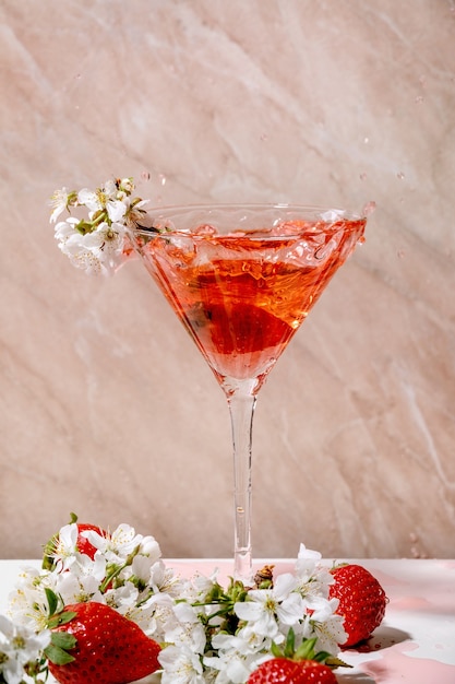 Plusk truskawkowego koktajlu alkoholowego lub bezalkoholowego w szkle ozdobionym kwitnącymi gałęziami wiśni na biało-różowej ścianie