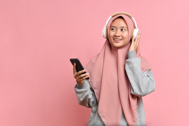 Pobudzona szczęśliwa muzułmanka tańczy radośnie, cieszy się ulubioną muzyką, nosi słuchawki stereo, patrzy na miejsce na kopię, odizolowana na różowym tle, wyraża pozytywny nastrój