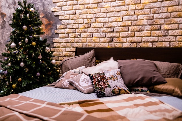 Pod ceglaną ścianą stoi duże podwójne łóżko z dywanikiem w kratkę, obok podwójnego łóżka stoi choinka. Wystrój noworoczny. Zdjęcie