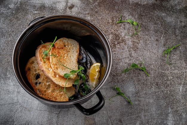 Podawanie dania z menu restauracji. Smażone małże z grzankami i cytryną na ciemnej patelni na szarym kamiennym stole