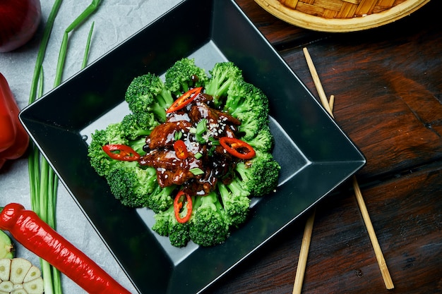 Zdjęcie podawanie świeżych gotowanych brokułów z kurczakiem w sosie teriyaki i papryczkami chili w czarnej misce na drewnianym stole. danie dietetyczne w stylu azjatyckim ze składnikami