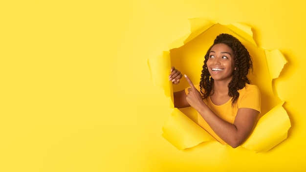 Podekscytowana Afroamerykanka wskazująca palcem na miejsce kopiowania, pozująca w rozdartej żółtej papierowej panoramie tła