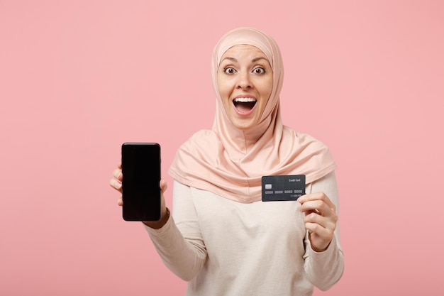 Podekscytowana arabska muzułmańska kobieta w jasnych ubraniach hidżab pozowanie na białym tle na różowym tle. Koncepcja życia religijnego ludzi. Makieta miejsca na kopię. Trzymaj telefon komórkowy z karty kredytowej bankowej puste pusty ekran.