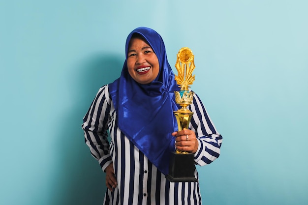 Podekscytowana Azjatka w średnim wieku w hidżabie trzyma złoty trofeum odizolowane na niebieskim tle