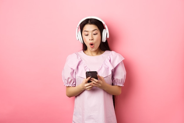 Podekscytowana Azjatycka Dziewczyna Patrząc Na Ekran Smartfona Rozbawiona, Powiedz Wow, Słuchając Muzyki W Słuchawkach Bezprzewodowych, Stojąc Na Różowym Tle.