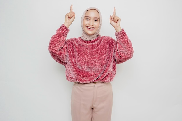 Podekscytowana azjatycka kobieta ubrana w różowy sweter i hidżab, wskazująca na miejsce kopiowania na niej, odizolowana białym tłem