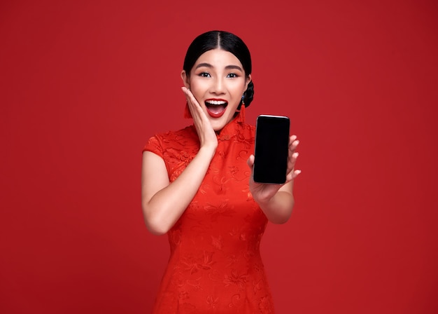 Podekscytowana Azjatycka kobieta ubrana w tradycyjny strój qipao cheongsam pokazujący telefon komórkowy na białym tle na czerwonej ścianie.