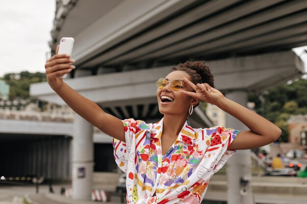 Podekscytowana ciemnoskóra brunetka z kręconymi włosami w kolorowej bluzce i stylowych pomarańczowych okularach przeciwsłonecznych pokazuje znak pokoju i robi sobie selfie na zewnątrz