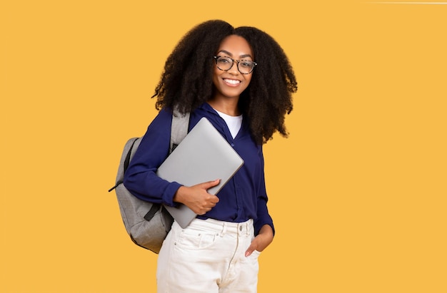 Zdjęcie podekscytowana czarna studentka z plecakiem trzymająca laptop i uśmiechająca się do kamery ustawionej przeciwko żółtemu