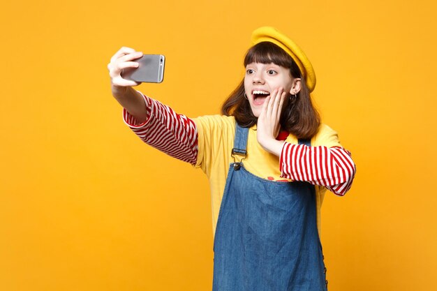 Podekscytowana dziewczyna nastolatek we francuskim berecie, dżinsowa sukienka robi selfie strzał na telefon komórkowy, kładąc rękę na policzku na białym tle na żółtym tle. Ludzie emocje, koncepcja stylu życia. Makieta miejsca na kopię.