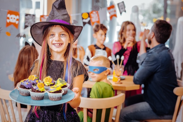 Podekscytowana dziewczyna. Promieniejąca szczęśliwa dziewczyna ubrana w kostium Halloween czuje się podekscytowany trzymając talerz z jasnymi babeczkami