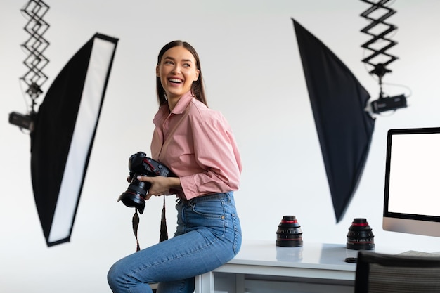 Podekscytowana fotografka siedząca na krawędzi stołu, trzymająca aparat DSLR i uśmiechnięte wnętrze studia fotograficznego
