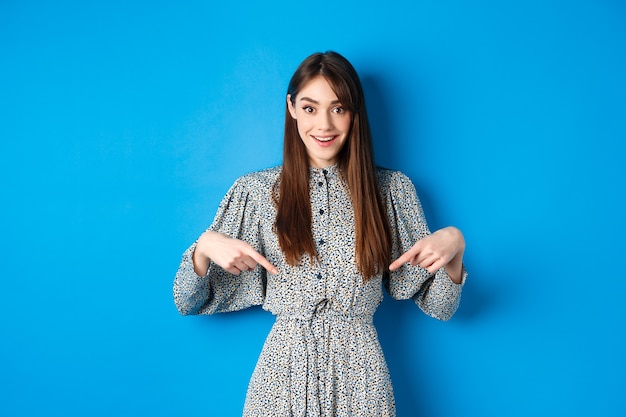 Podekscytowana kaukaska kobieta w sukience wskazująca palcami w dół na logo, pokazująca fantastyczne wiadomości i uśmiechnięta, stojąca na niebiesko.