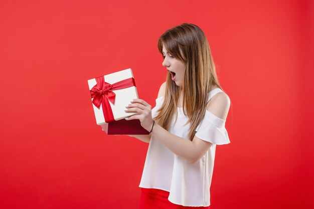 Podekscytowana kobieta otwierając pudełko na sobie białą bluzkę na białym tle na czerwonej ścianie