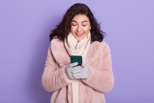 Podekscytowana kobieta trzyma smartfon, czyta wiadomości z serwisów społecznościowych, nosi stylowy różowy płaszcz ze sztucznego futra, szalik i rękawiczki