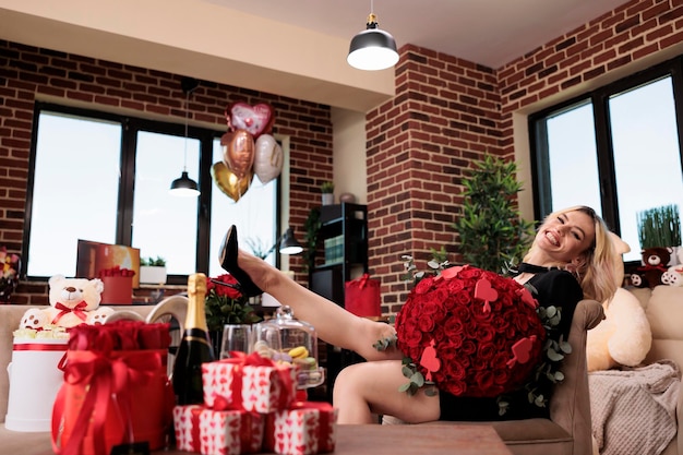 Podekscytowana kobieta trzymająca czerwone róże, śmiejąca się w pokoju wypełnionym luksusowymi prezentami, patrząca w kamerę. Szczęśliwa blondynka z drogim bukiet kwiatów siedzi, podnosząc portret nogi.