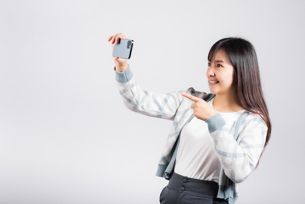 Podekscytowana kobieta trzymająca smartfona do robienia zdjęć selfie przednią kamerą studio na białym tle, szczęśliwa młoda kobieta uśmiechająca się, robiąca zdjęcia telefonem komórkowym na telefonie komórkowym