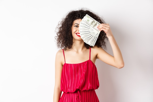 Podekscytowana kobieta w czerwonej sukience wygrywająca pieniądze, pokazująca dolary i uśmiechnięta szczęśliwa, stojąca na białym tle.