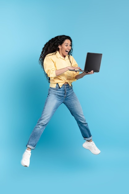 Podekscytowana kobieta z Bliskiego Wschodu trzymająca laptopa skaczącego na niebieskim tle