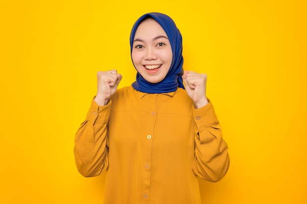 Podekscytowana młoda azjatycka muzułmanka ubrana na pomarańczowo świętuje sukces z uniesionymi pięściami na żółtym tle