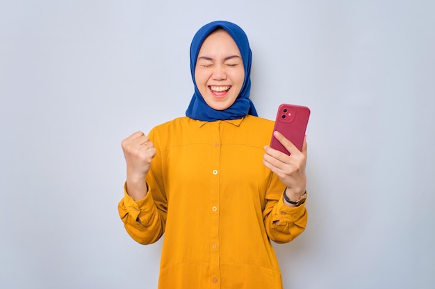 Podekscytowana młoda azjatycka muzułmanka ubrana w pomarańczową koszulę, korzystająca z telefonu komórkowego, świętująca sukces otrzymywania dobrych wiadomości na białym tle na białym tle