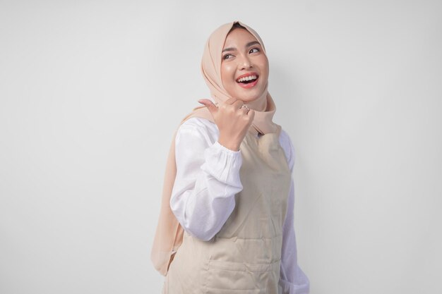 Podekscytowana młoda azjatycka muzułmańska kobieta w hidżabie i kremowym fartuchu wskazująca na pustą przestrzeń do kopiowania po lewej i prawej stronie koncepcji reklamy Ramadanu